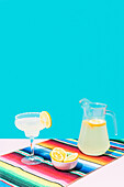 Glas und Krug mit kalter Limonade mit frischen Zitronen auf bunter mexikanischer Teppichdecke auf blauem Hintergrund