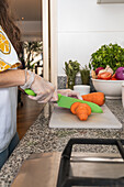 Crop anonymous female in gloves cutting slices of ripe orange carrot on chopping board with salad while preparing healthy vegetarian dish on counter with assorted vegetables in kitchen