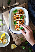 From above hand with cauliflower snack tacos in bowl and lemon pieces in plate on wooden table