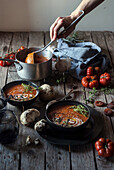 Crop person holding soup ladle and serving creamy tomato soup with seeds on table with dried apricots and bread