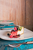 Leckerer Käsekuchen mit roter Marmelade, reifen Himbeeren, Blaubeeren, frischen Minzblättern und Puderzucker auf weißem Keramikteller im Restaurant