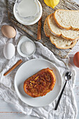 Blick von oben auf einen Teller mit appetitlichem spanischem Torrija-Brot, das mit rohen Eiern und Zucker auf dem Tisch liegt, neben einem hölzernen Schneidebrett mit einem Krug frischer Milch und Brotscheiben