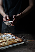 Crop shot of woman covering freshly baked frangipane apple pie with powdered sugar on wooden table