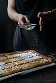 Crop shot of woman covering freshly baked frangipane apple pie with powdered sugar on wooden table