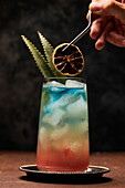 Crop unrecognizable person preparing glass of rainbow paradise colorful cocktail garnished with orange slices and leaves on slate tray on table