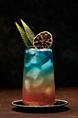Glas Regenbogenparadies mit buntem Cocktail, garniert mit Orangenscheiben und Blättern auf einem Schiefertablett auf dem Tisch