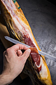 top view of Crop unrecognizable cook cutting slice of tasty dry cured Spanish jamon on black background