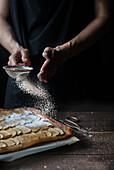 Crop shot of woman covering freshly baked frangipane apple pie with powdered sugar on wooden table