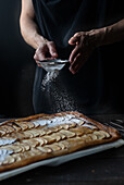 Crop shot of woman covering freshly baked frangipane apple pie with powdered sugar on wooden table