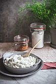 Keramikschale mit leckerem Frischkäse und Holzlöffel auf Marmortisch neben Gewürzen und Kräutern