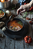 Crop person holding soup ladle and serving creamy tomato soup with seeds on table with dried apricots and bread