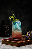 Glass of rainbow paradise colorful cocktail garnished with slice of orange and flowers on old book board on table