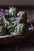 Closeup from above view of green artichokes laid in wicker basket with bunch of little purple flowers