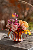 Blumenstrauss aus Rosen (Rosa) und Schneebeere (Symphoricarpos) 'Amethyst' in Vase auf Gartenbank