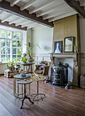 Living room with antique wood-burning stove, plants and wooden floor, large window and wooden beamed ceiling