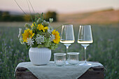 Spätsommerstrauß aus Hortensien (Hydrangea) und Sonnenblumen mit Gläsern auf Gartentisch bei Sonnenuntergang