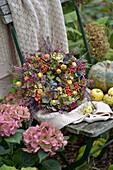 Bunter Herbstkranz aus Hortensien (Hydrangea), Samenkapseln, Heidekraut, Zieräpfeln und Vogelbeeren auf rustikalem Gartenstuhl