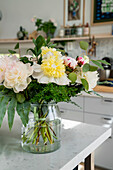 Fresh bouquet of flowers with peonies and ferns in glass vase on kitchen worktop