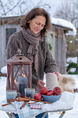 Frau mit Laterne und Keramik Krug, Tassen, Äpfel, Zimtstangen in winterlichem Garten und Gartenhütte