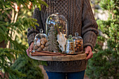 Frau mit Glasglocken gefüllt mit Glitzertanne, Walnüssen, Zimtstangen, Hirschgeweih und Keksen auf Holzschscheibe im Garten
