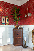 Antique chest with palm tree in a pot and wall candle holder in front of red wallpaper