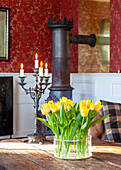 Yellow tulips (Tulipa) in glass vase on wooden table, antique wood-burning stove in the background