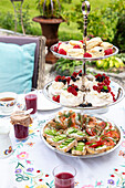 Etagere mit belegten Broten, Mini-Pavlovas und Scones im Garten, Tischdecke mit Blumenmuster