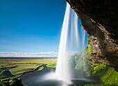 Blick auf den Seljalandsfoss-Wasserfall, der von einer Klippe herabstürzt, mit blauem Himmel und Landschaft im Hintergrund, Südliche Region, Island