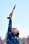 Kleiner Junge hält eine gelbe und blaue Spielzeugrakete in der Luft vor blauem Himmel