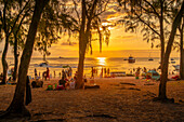 Blick auf Bäume und Menschen am öffentlichen Strand von Mon Choisy bei Sonnenuntergang, Mauritius, Indischer Ozean, Afrika