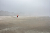 Frau in orangefarbener Jacke steht am nebligen Strand