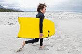 Junge (10-11), der am Strand läuft und ein Bodyboard trägt