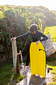 Junge (10-11) mit Surfbrett an einem Wasserhahn im Freien am Kammabaai Beach