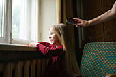 Mädchen (4-5) schaut durch das Fenster, während die Mutter ihr die Haare bürstet