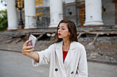 Porträt einer jungen Frau, die ein Selfie mit ihrem Smartphone macht, während sie vor einem Gebäude steht