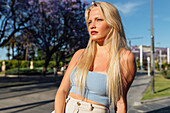 Charming female with blond hair and in trendy summer clothes standing in city and looking away