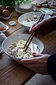 Ausschnitt einer nicht erkennbaren Person, die frisch gekochte Ramen-Nudeln mit Tofu, Eiern und Gemüse mit Stäbchen auf einem Holztisch zubereitet