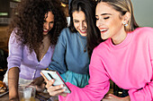 Smiling young female friends wearing casual clothes browsing mobile phones while gathering for lunch in restaurant