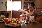 Fokussierte ethnische Hausfrau wiegt frische Tomaten in einem Glaskrug auf einer Küchenwaage, während sie zu Hause Essen kocht