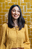 Young friendly female in yellow wear looking at camera with toothy smile against bright wall