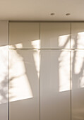 Einfacher weißer Holzschrank mit geschlossenen Türen in einer modernen Küche mit Sonnenlicht und Schatten von Bäumen