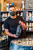 Seriöser bärtiger Barkeeper mit dunklem Haar in schwarzer Schürze steht am Tresen und zeigt eine Flasche Rotwein, während er im Restaurant arbeitet