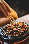 Hands of anonymous woman using chopsticks to eat tasty beef with vegetables placed on wooden plate