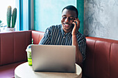 Content African American male entrepreneur speaking on cellphone against table with netbook and smoothie in restaurant