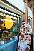 Cool male in sunglasses with healthy drink to go looking away while leaning on urban signboard with photo of food