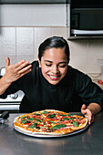 Young positive chef woman in black uniform smelling aroma of tasty fresh cooked pizza placed on table