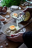Abgeschnittene unerkennbare Person, die frisch gekochte Ramen-Nudeln mit Tofu, Eiern und Gemüse mit einer Flasche Brühe auf einem Holztisch zubereitet
