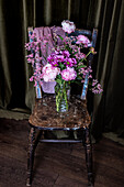 Strauß frischer bunter Pfingstrosen und Chrysanthemen in einer Glasvase auf einem verwitterten Holzstuhl neben Vorhängen in einem hellen Raum