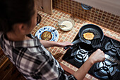 From above side view of focused young Asian female in casual clothes cooking yummy pancakes using spatula while standing in kitchen