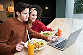 Fröhliches multiethnisches Paar isst gesundes Frühstück im Restaurant und surft auf dem Laptop